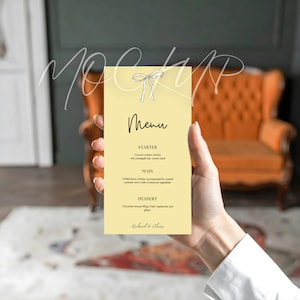 Op de afbeelding: Een rechthoekige menukaart met een lichtgele achtergrond wordt in de hand van een persoon gehouden. Het menu heeft een strik-afbeelding bovenaan en het woord "Menu" in cursief. Het menu bevat een voorgerecht, hoofdgerecht en dessert.