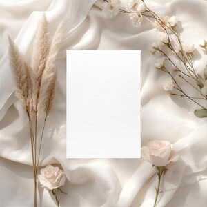 May include: A blank white card on a cream-colored silk fabric, surrounded by beige pampas grass, white flowers, and pink roses. The composition is arranged in a flat lay style, creating a soft and elegant aesthetic.