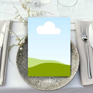 May include: A place setting with a speckled gray plate, silverware, and a folded napkin. A card with a landscape design featuring a blue sky, white cloud, and green hills sits on the plate. Delicate white flowers and a clear glass are also visible.