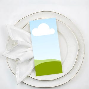 Peut inclure: Une carte de place rectangulaire avec un ciel bleu et un nuage, et des collines vertes en bas, repose sur une assiette blanche avec un bord doré. Une serviette blanche est pliée en forme décorative.