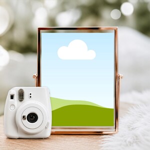 May include: A white instant camera sits on a wooden surface next to a rose gold framed picture. The picture displays a blue sky with a white cloud and green hills. The camera has a lens and flash.