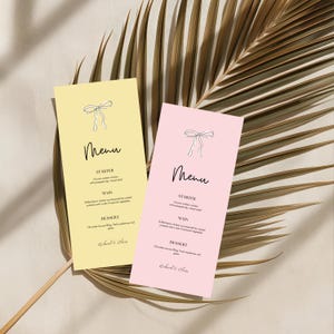 May include: Two rectangular menus, one pale yellow and one pink, with a bow illustration at the top. The word "Menu" is written in elegant script. The menus are placed on a dried palm leaf.
