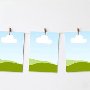May include: Three landscape art prints hanging from a string with wooden clips. Each print features a blue sky with a white cloud and a green rolling hill. The prints are displayed against a white background.