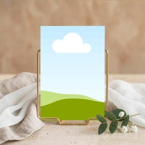 May include: A gold metal picture frame holding a landscape art print with a blue sky, white cloud, and green hills. The frame is on a beige surface with a white cloth and greenery.