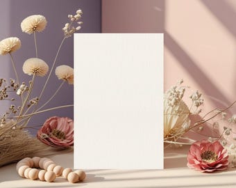 May include: A blank, off-white rectangular card stands upright on a light-colored table, surrounded by dried flowers and a wooden bead garland. The background features a soft color palette of pink and purple.