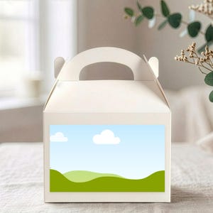 Könnte beinhalten: Eine weiße Giebelbox mit Griff, mit einem Landschaftsdesign auf der Vorderseite. Das Design zeigt einen blauen Himmel mit weißen Wolken und grünen Hügeln. Die Box steht auf einer weißen Oberfläche.