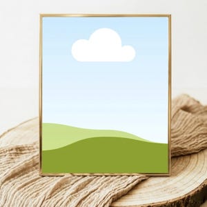 May include: A framed art print featuring a minimalist landscape scene. The artwork depicts a blue sky with a white cloud above rolling green hills. The frame is gold-colored, and the print is displayed on a wooden surface with a tan cloth.