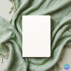 May include: A blank, white notebook with rounded corners rests on a soft, sage green fabric with fringe. The notebook's cover has a textured appearance. The fabric is draped, creating folds and shadows.