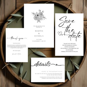 May include: A collection of white wedding stationery cards with black text. The cards include a floral invitation, a save-the-date card, a thank you card, and a details card. The cards are arranged on a wooden tray with greenery.