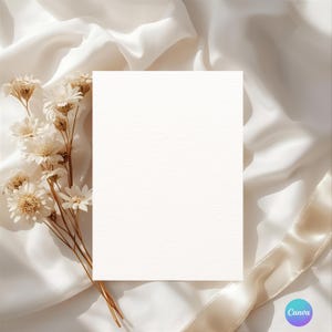Op de afbeelding: Een blanco, getextureerde witte kaart is gecentreerd op een achtergrond van ivoorkleurige zijden stof. Gedroogde witte bloemen en een crèmekleurig lint zijn rond de kaart gerangschikt, wat een zachte, elegante esthetiek creëert. Het Canva-logo staat rechtsonder.
