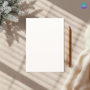 Puede incluir: Un papel blanco en blanco con una superficie texturizada, un lápiz de madera y una tela suave de color blanquecino. La escena está ambientada sobre una superficie beige con sombras de una ventana y una rama de pino escarchada.