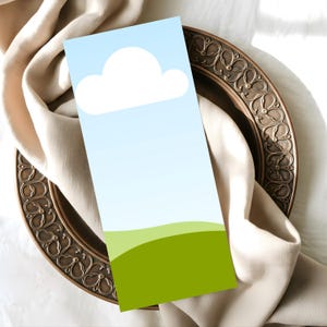 Puede incluir: Una tarjeta rectangular con un diseño de cielo y nube, con un fondo degradado azul y una nube blanca. La parte inferior de la tarjeta tiene un diseño de colina verde. La tarjeta está colocada sobre un plato decorativo de bronce y una tela beige.