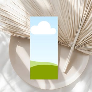 May include: A rectangular bookmark with a sky and landscape design. The top portion features a white cloud against a light blue gradient, transitioning to a green field at the bottom. The bookmark is set on a white plate with a dried palm leaf.