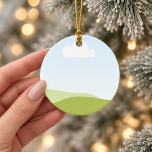 May include: A round ceramic ornament featuring a minimalist landscape design with a light blue sky, a single white cloud, and rolling green hills. The ornament is suspended by a thin gold cord, creating a simple and clean aesthetic.