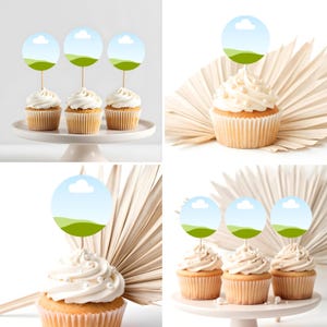 May include: Four cupcakes with white frosting and decorative toppers featuring a blue sky and green landscape design. The cupcakes are on a white cake stand, perfect for a party or celebration.