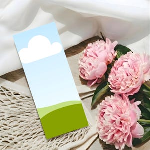 May include: A bookmark with a sky and cloud design, featuring a gradient blue background, a white cloud, and a green hill. The bookmark is placed near pink peonies and a white mesh bag.
