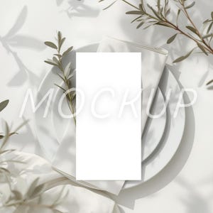 May include: A white rectangular card with the word "MOCKUP" in light gray text, placed on a white plate with a folded white napkin. Olive branches and their shadows add a natural touch to the composition.
