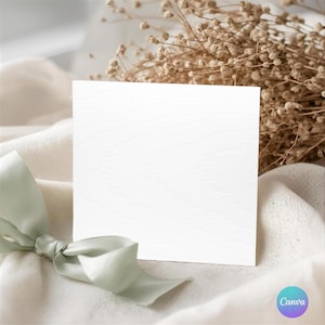 Peut inclure: Une carte carrée blanche vierge avec une texture de grain de bois, posée sur un tissu crème doux. Un ruban vert clair est noué en bas à gauche. Des fleurs séchées de couleur beige sont en arrière-plan.