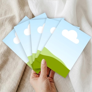 May include: A hand holding a fanned stack of postcards. Each postcard features a blue sky with white clouds and a green grassy hill. The postcards are arranged on a beige and white fabric background.