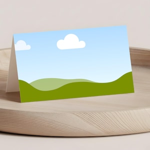 May include: A folded greeting card featuring a landscape design with a blue sky, white clouds, and green rolling hills. The card is displayed on a light wooden tray.