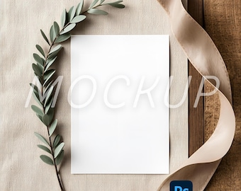 May include: A blank white card with the word "MOCKUP" overlaid in a light color, with a sprig of green leaves and a beige ribbon. The background is a mix of beige fabric and wood.