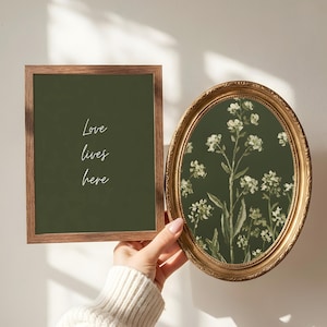 Può includere: Due opere d'arte incorniciate. Una è rettangolare con una cornice in legno e il testo "Love lives here" in bianco. L'altra è una cornice ovale con finitura dorata, con una stampa floreale di fiori bianchi.