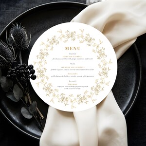 Puede incluir: Una tarjeta de menú redonda con detalles florales dorados y la palabra "MENU" en la parte superior. El menú enumera entrantes, platos principales y postres. La tarjeta está sobre un plato negro con una servilleta blanca y flores negras decorativas.