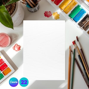 May include: A blank white textured paper is centered on a white surface, surrounded by art supplies. Paintbrushes, watercolor paints, a potted plant, and the text "Canva 22" are visible, suggesting a creative workspace.