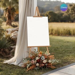 Puede incluir: Un caballete de madera con un letrero blanco en blanco, decorado con un arreglo floral. Las flores son en tonos de rosa, melocotón y burdeos. El caballete está instalado al aire libre sobre hierba.