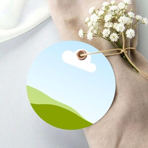 Peut inclure: Une étiquette cadeau ronde avec un ciel bleu et un design de nuage, avec un paysage vert en bas. L'étiquette a un œillet doré et est attachée avec de la ficelle. Un petit bouquet de fleurs blanches est attaché à une serviette beige.