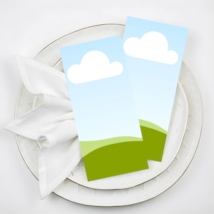 May include: Two rectangular place cards with a sky and cloud design, on a white plate with a gold rim. A white napkin is folded decoratively. The place cards feature a blue sky, white cloud, and green hills.