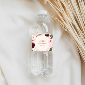 K&ouml;nnte beinhalten: Eine durchsichtige Wasserflasche aus Kunststoff mit wei&szlig;em Verschluss und einem rechteckigen Etikett. Das Etikett zeigt ein florales Design mit burgunderroten und hellrosa Rosen, die einen zentralen Kreis mit dem Text Mia's Baby Shower und dem Datum 12.07.2025 umgeben.