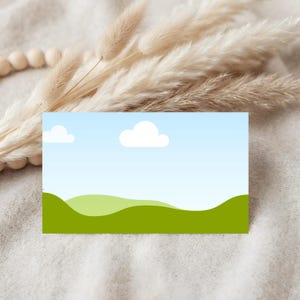 May include: A rectangular card featuring a landscape design with a blue sky, white clouds, and green rolling hills. The card is set against a neutral background with dried ornamental grass and wooden beads.
