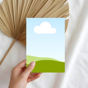 Puede incluir: Una mano sosteniendo una tarjeta de felicitación con un diseño de paisaje minimalista. La tarjeta presenta una nube blanca contra un cielo azul degradado y colinas verdes. Una hoja de palma seca y tela blanca están en el fondo.