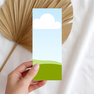 May include: A rectangular bookmark featuring a sky and landscape design. The top portion depicts a light blue sky with a white cloud. The bottom has green rolling hills. The bookmark is held by a hand.
