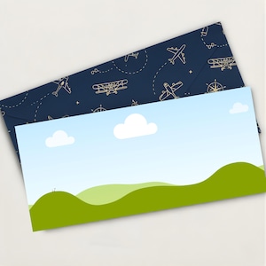 May include: Two-piece stationery set. The top envelope is navy blue with gold airplane and compass illustrations. The bottom card features a landscape with green hills, blue sky, and white clouds. The stationery set is on a white background.