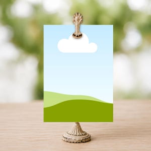 May include: A rectangular card featuring a light blue sky, a single white cloud, and rolling green hills, held upright by a vintage-style metal stand with a decorative clip at the top. The card is positioned vertically and displays a simple landscape.