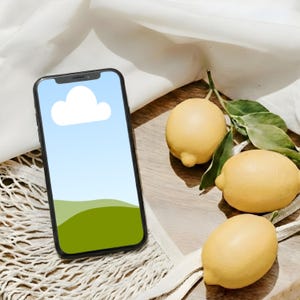 以下が含まれることがあります： 空と雲のグラフィックが表示された画面を持つ黒いスマートフォンが、木製のトレイに置かれた3つの黄色のレモンの隣にあります。白い布と白いメッシュバッグがシーンを完成させています。