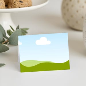 Puede incluir: Una tarjeta plegada con un diseño de paisaje que presenta un cielo azul, una nube blanca y colinas verdes. La tarjeta es blanca por dentro y probablemente se utiliza para un saludo o una invitación. La tarjeta está sobre una superficie blanca.