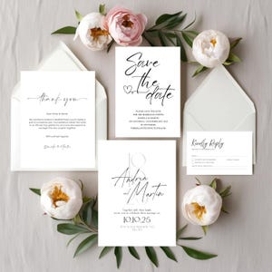 May include: A collection of white wedding stationery with black calligraphy. The set includes a "Save the Date" card, a "Thank You" card, an invitation, and a reply card. The cards are surrounded by white envelopes and pink and white flowers.