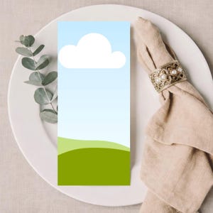 May include: A rectangular card with a sky and cloud design, placed on a white plate. The card features a blue gradient background, a white cloud, and green hills. A beige napkin with a decorative gold ring is on the plate.