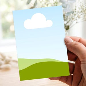 Op de afbeelding: Een rechthoekige kaart met een minimalistisch landschapsontwerp. Het toont een lichtblauwe lucht met een witte wolk en groene heuvels. De kaart wordt door een hand vastgehouden.