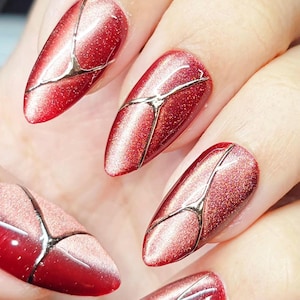 May include: Close-up of a hand with five almond-shaped nails. The nails are painted a shimmering red with a silver geometric design. The design appears to be a series of intersecting lines across each nail.