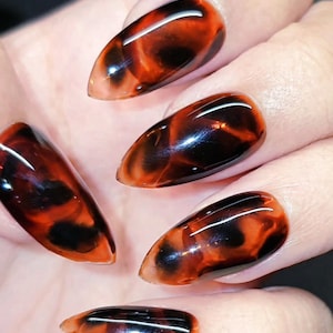 May include: Close-up of a hand displaying five almond-shaped acrylic nails. The nails showcase a tortoiseshell pattern, combining translucent amber and dark brown swirls. The nails are glossy and well-manicured, with a smooth finish.