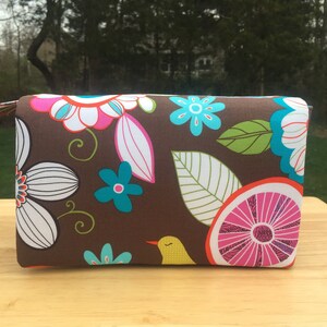 K&ouml;nnte beinhalten: Eine rechteckige braune Stofftasche mit einem farbenfrohen Blumenmuster. Das Design umfasst wei&szlig;e, rosa und t&uuml;rkisfarbene Blumen, gr&uuml;ne Bl&auml;tter und einen gelben Vogel. Die Tasche hat einen passenden Rei&szlig;verschluss.