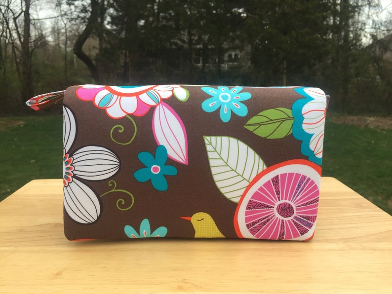 K&ouml;nnte beinhalten: Eine rechteckige braune Stofftasche mit einem farbenfrohen Blumenmuster. Das Design umfasst wei&szlig;e, rosa und t&uuml;rkisfarbene Blumen, gr&uuml;ne Bl&auml;tter und einen gelben Vogel. Die Tasche hat einen passenden Rei&szlig;verschluss.