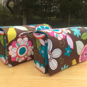 K&ouml;nnte beinhalten: Zwei braune Stoffbeutel mit einem farbenfrohen Blumen- und Vogelmuster. Das Design umfasst gro&szlig;e wei&szlig;e, rosa und t&uuml;rkisfarbene Blumen, gr&uuml;ne Bl&auml;tter und gelbe V&ouml;gel. Jeder Beutel hat einen t&uuml;rkisfarbenen Rei&szlig;verschluss und einen kleinen Griff.