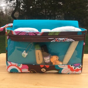 K&ouml;nnte beinhalten: Eine rechteckige Kosmetiktasche mit transparenten Seiten, einem t&uuml;rkisfarbenen Innenraum und einem bunten Blumenmuster. Die Tasche enth&auml;lt verschiedene Gegenst&auml;nde, darunter H&auml;ndedesinfektionsmittel, eine Sicherheitsnadel und Wattepads. Die Tasche steht auf einer Holzoberfl&auml;che.