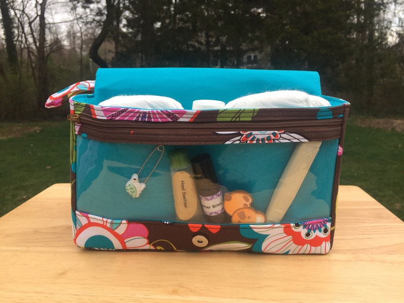 K&ouml;nnte beinhalten: Eine rechteckige Kosmetiktasche mit transparenten Seiten, einem t&uuml;rkisfarbenen Innenraum und einem bunten Blumenmuster. Die Tasche enth&auml;lt verschiedene Gegenst&auml;nde, darunter H&auml;ndedesinfektionsmittel, eine Sicherheitsnadel und Wattepads. Die Tasche steht auf einer Holzoberfl&auml;che.