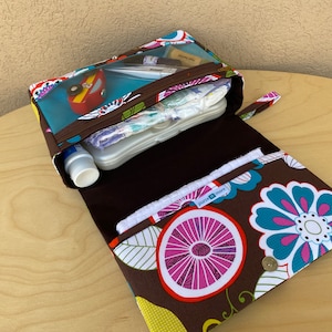 K&ouml;nnte beinhalten: Eine braune Wickelclutch mit floralem Muster, ge&ouml;ffnet, um Windeln, Feuchtt&uuml;cher und andere Babyutensilien zu zeigen. Die Clutch hat ein transparentes Rei&szlig;verschlussfach und einen Druckknopfverschluss. Das Innere ist einfarbig braun.